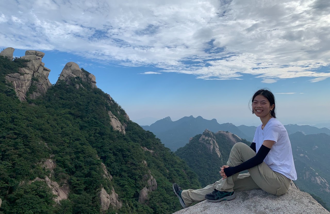 Wanchun Shen sitting on a granite outcrop, with forested mountain peaks behind her.